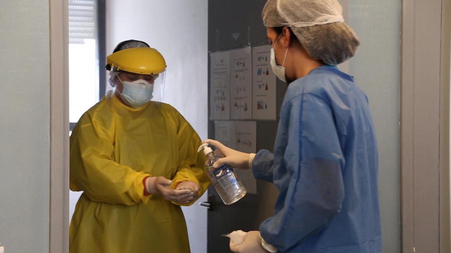Dos médicas con túnicas descartables y tapabocas, una pasándole alcohol en gel a las manos de la otra.